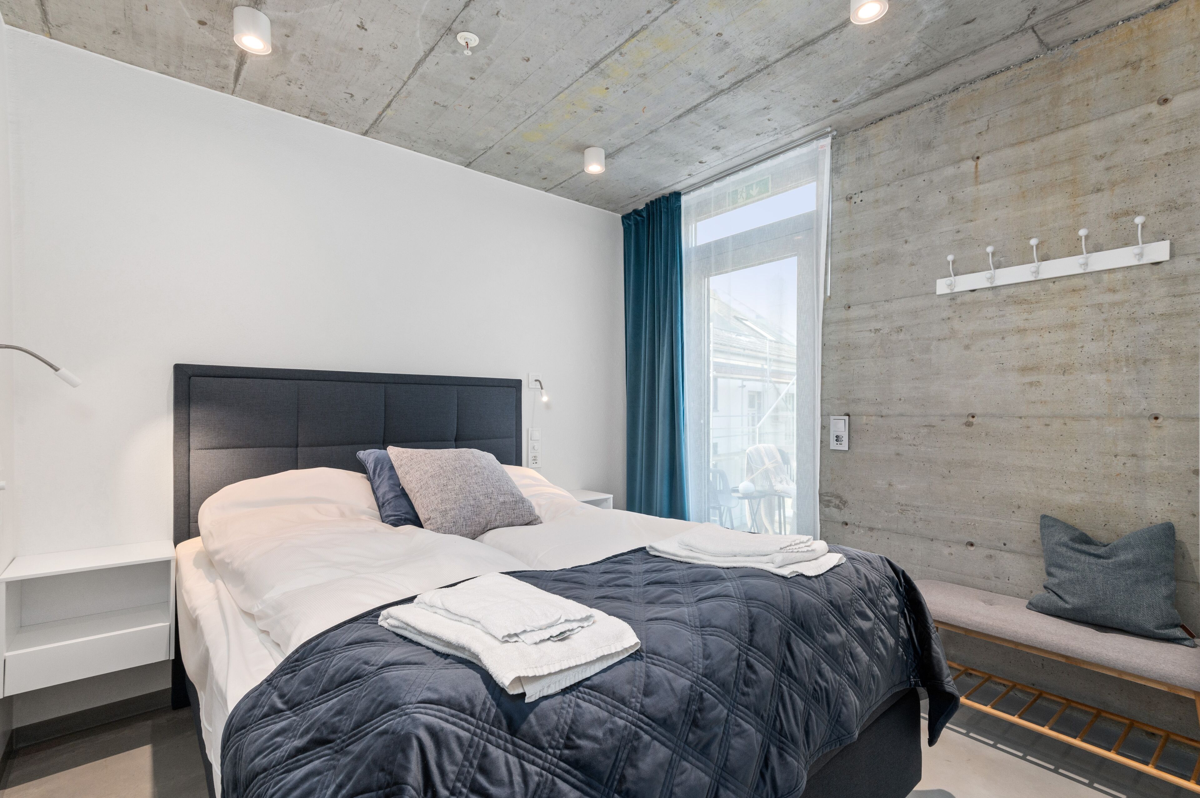 Un dormitorio moderno con una cama grande, una banca, un estante en la pared y una ventana con cortinas.