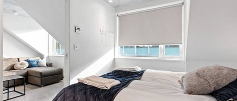 A modern bedroom with a bed, a window with blinds, a sofa, and a staircase leading to an upper level.