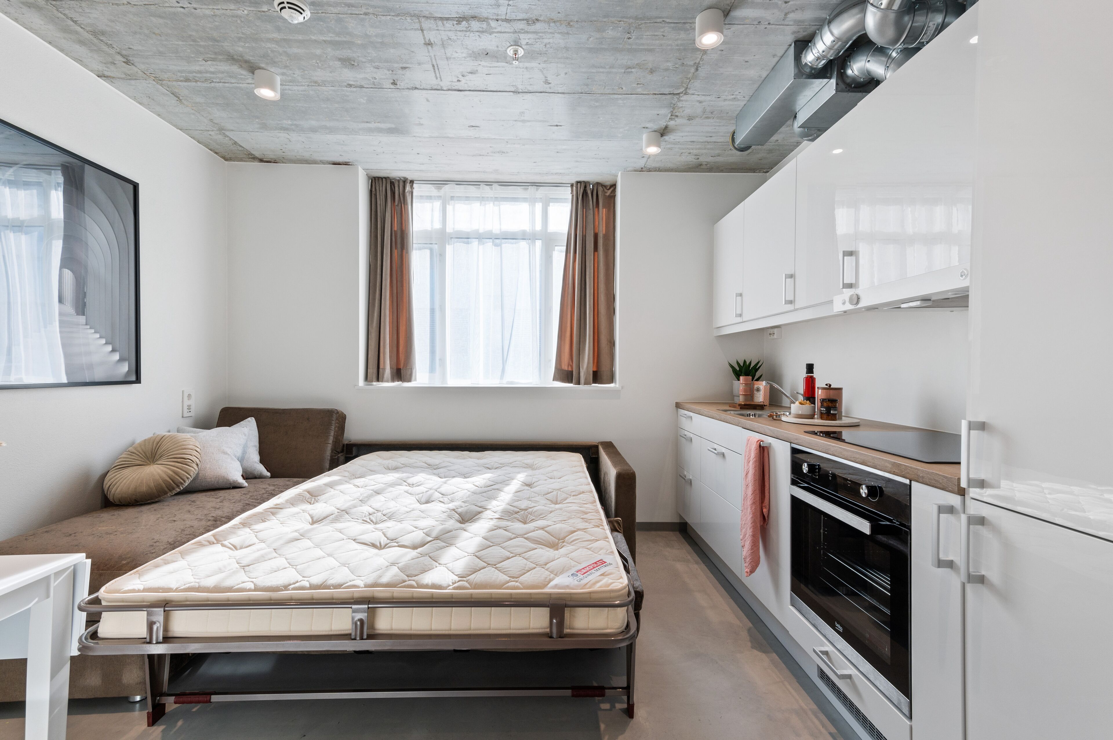 A compact bedroom with a bed, a small kitchenette, and a window with curtains.