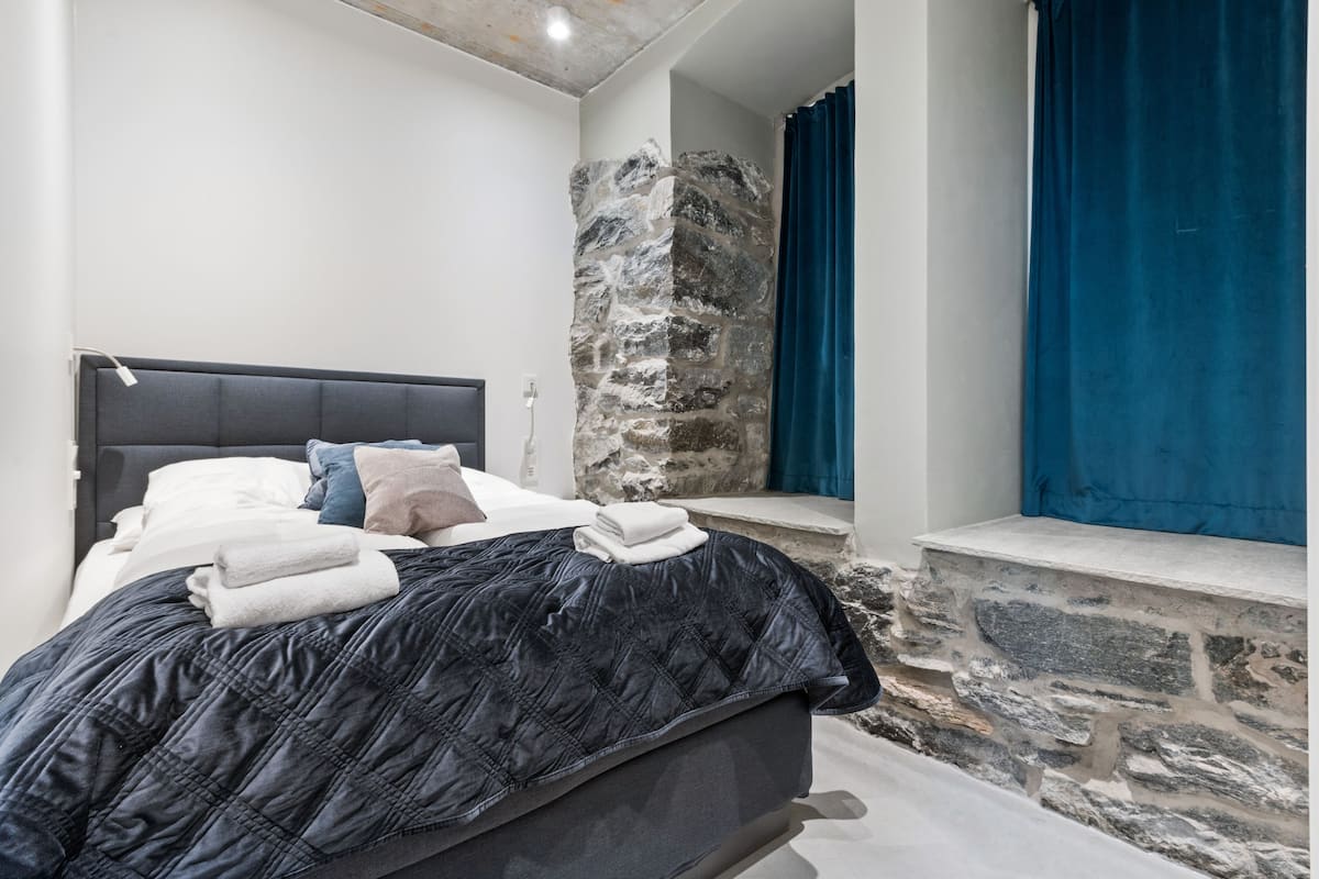 Une chambre moderne avec un mur en pierre, un lit recouvert d’une couverture sombre et des rideaux bleus.