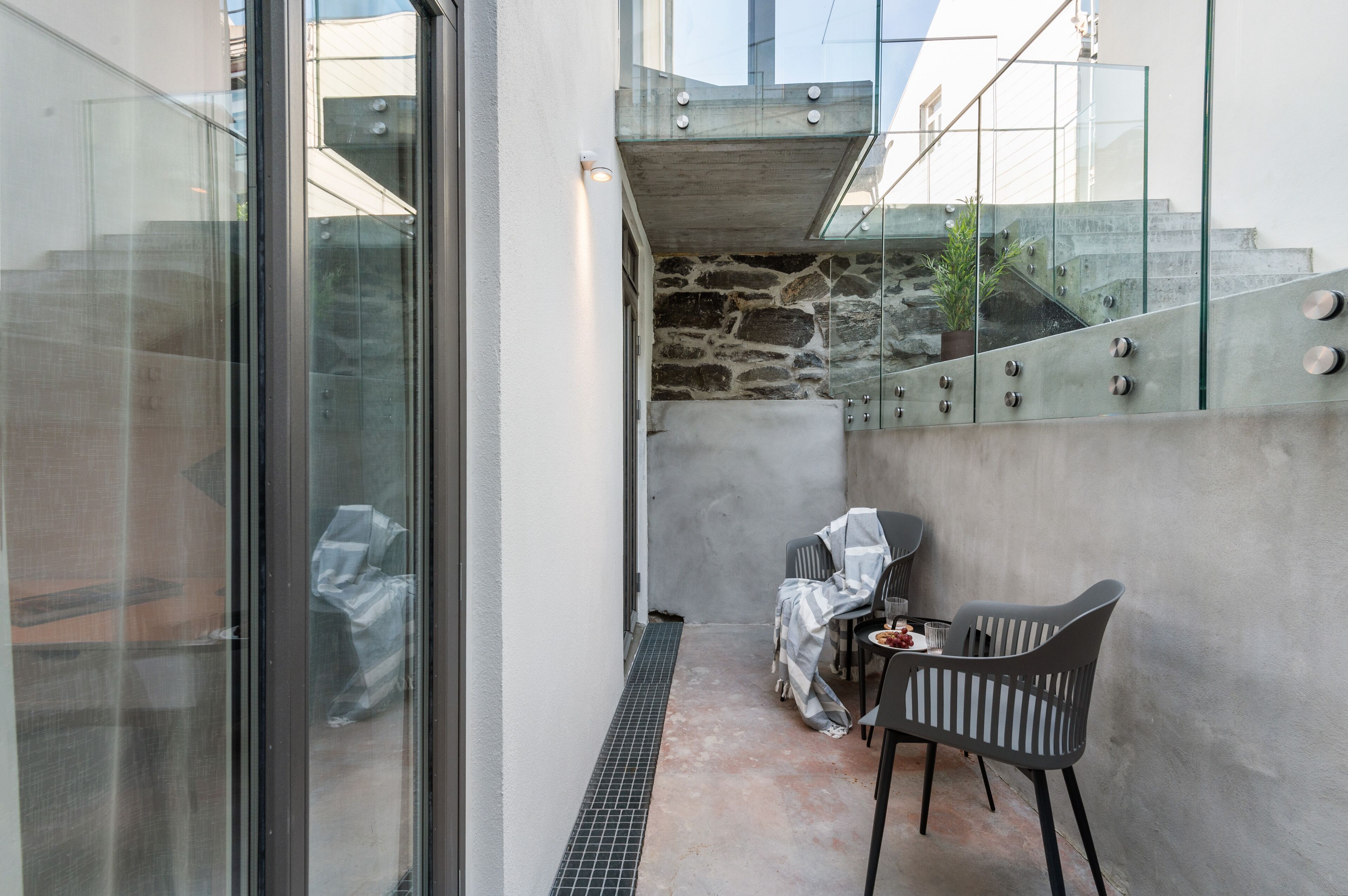 Un espacio exterior moderno con zona de estar, pared de vidrio, pared de piedra y escalera de metal.