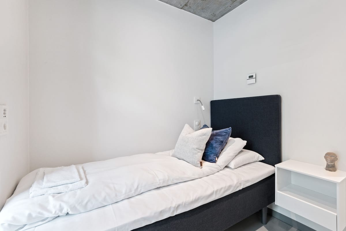 Une chambre à coucher moderne avec un lit simple, du linge de lit blanc, une tête de lit sombre et une table de chevet blanche ornée d’une petite sculpture.