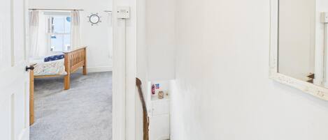 Un couloir avec un escalier, un lit avec une tête de lit en bois, un mur blanc avec un miroir et une fenêtre avec des rideaux.