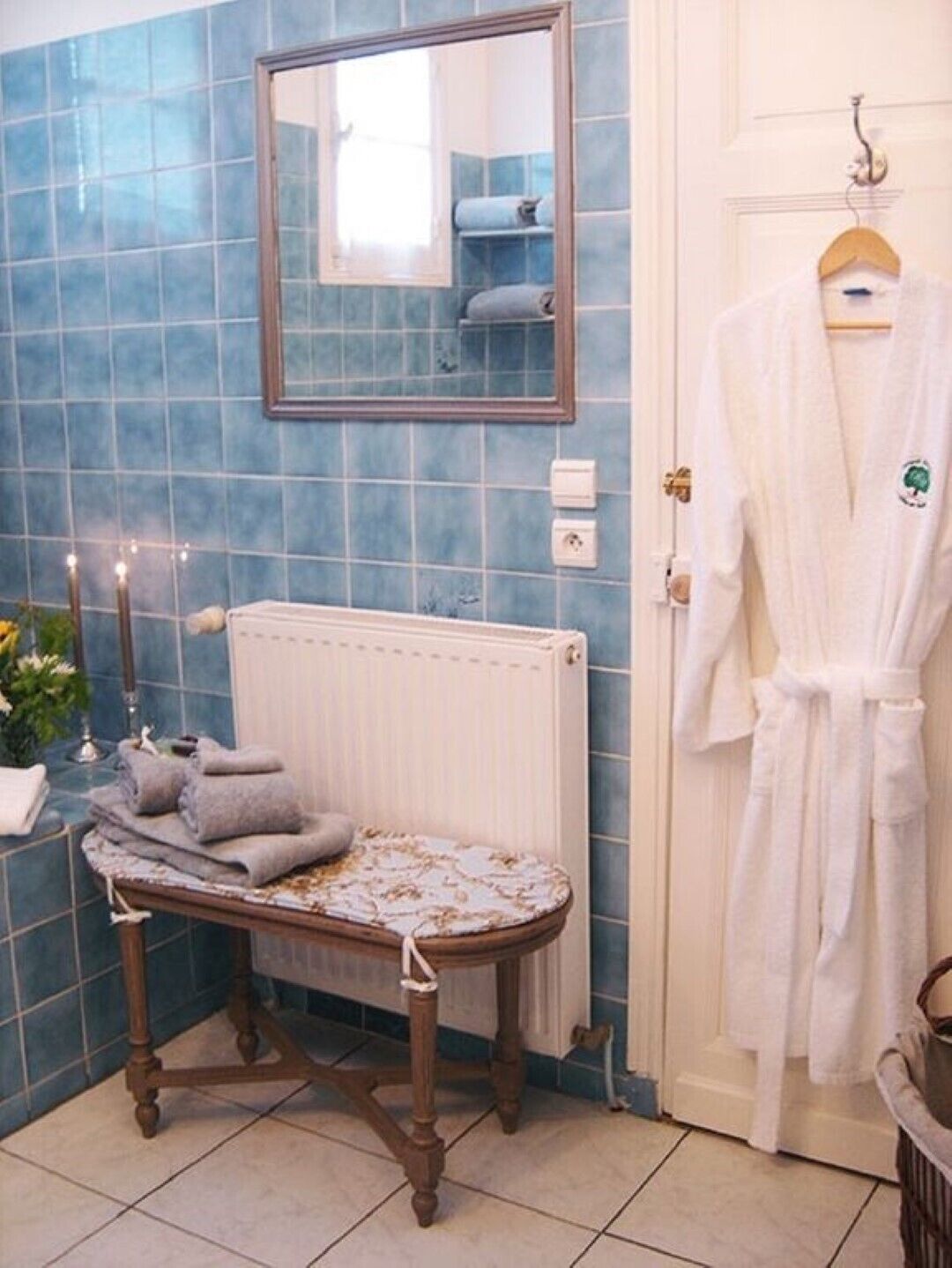 Une salle de bain avec des carreaux bleus, un banc en bois, un radiateur, une robe blanche sur un cintre et un miroir.