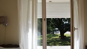 A room with a bed, a window with white blinds, and a view of a tree and lawn outside. - Rural country retreat with private heated pool and driving range! (Verteuil-D'agenais)