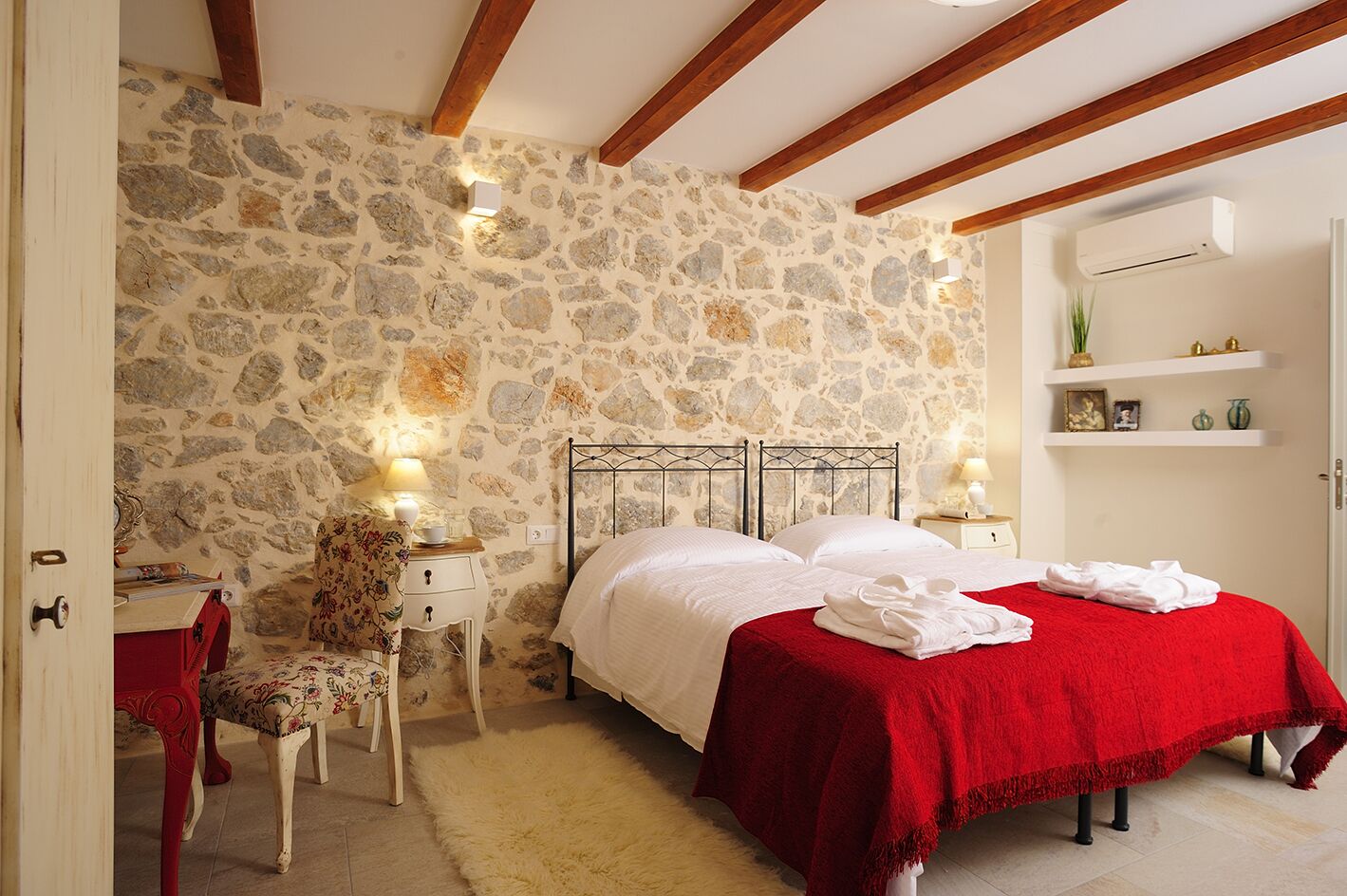 A bedroom with a stone wall, a bed with a red bedspread, a nightstand, a chair, and a small shelf with decorative items.