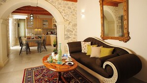 A cozy living room with a brown sofa, a coffee table, and a patterned rug. An archway leads to a kitchen with wooden cabinets and a stone wall. - Hawk Villa, Timeless Village Escape with Pool (Gerakari, Rethymno)