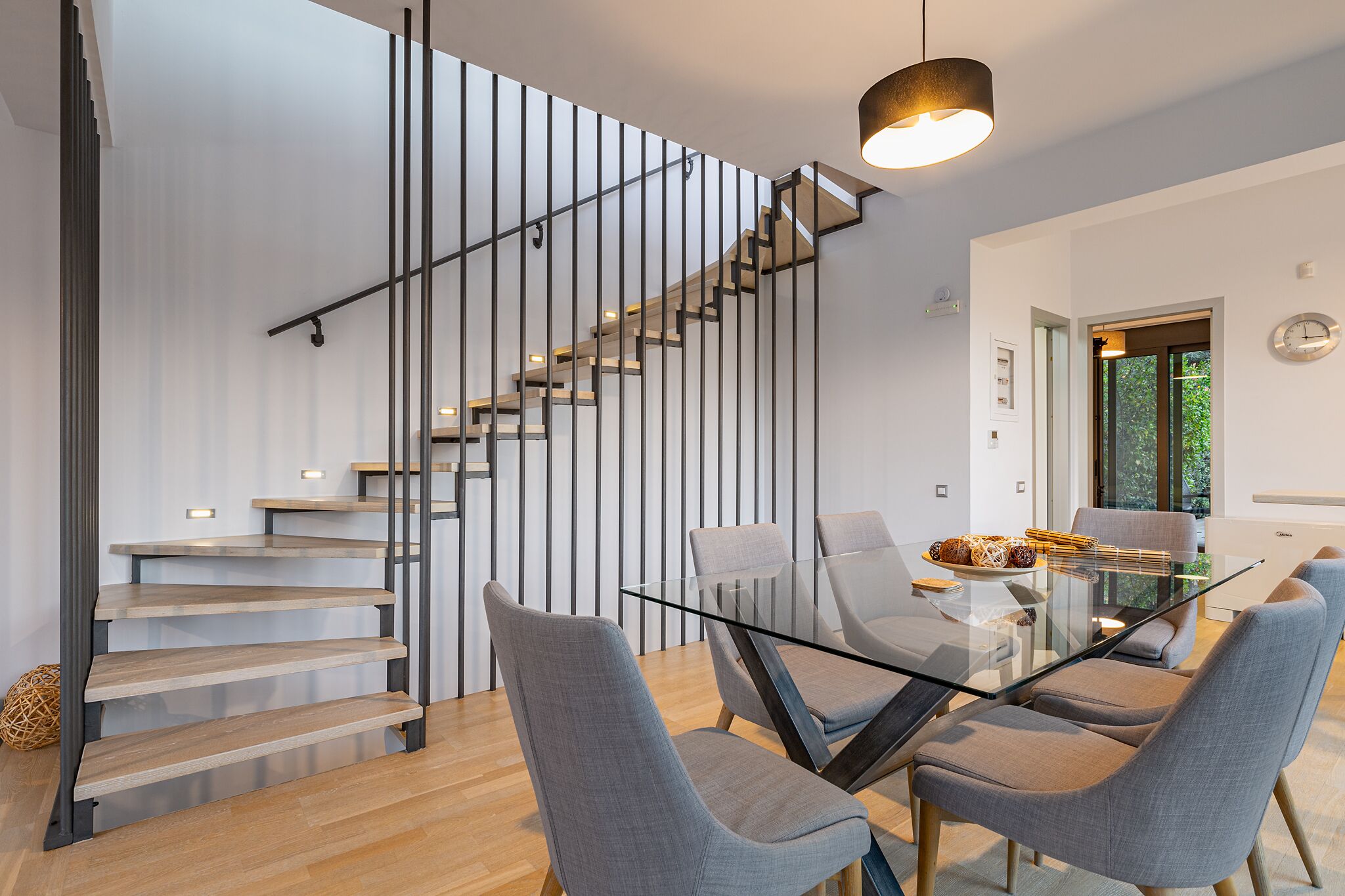 A modern interior with a glass-top dining table, grey upholstered chairs, a staircase with wooden steps, and a pendant light.