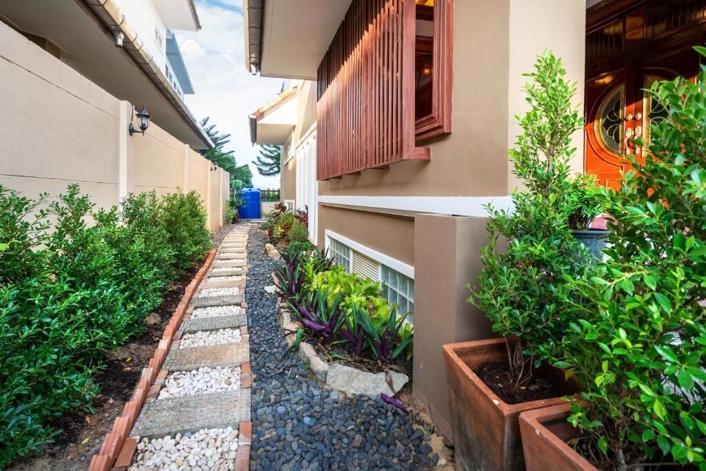 Un étroit sentier bordé de plantes en pot et un bâtiment à la façade beige et aux volets en bois.