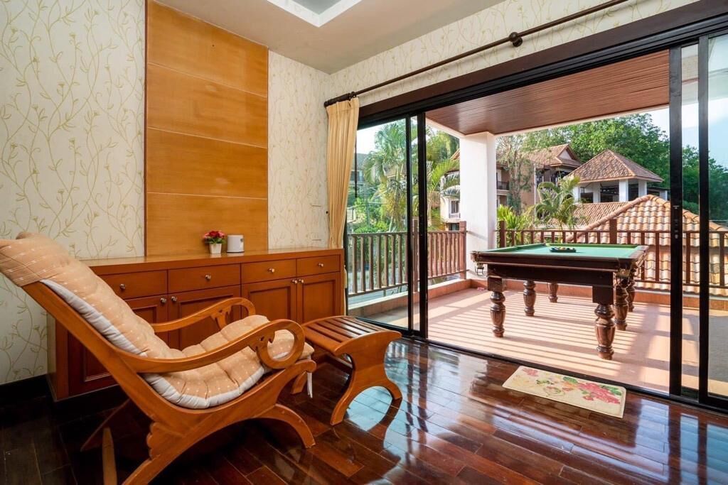 A room with a wooden sofa, a pool table, and a view of a balcony with a wooden deck and a building in the background.