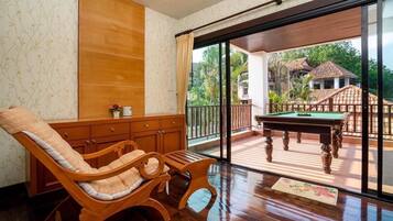 A room with a wooden sofa, a pool table, and a view of a balcony with a wooden deck and a building in the background.