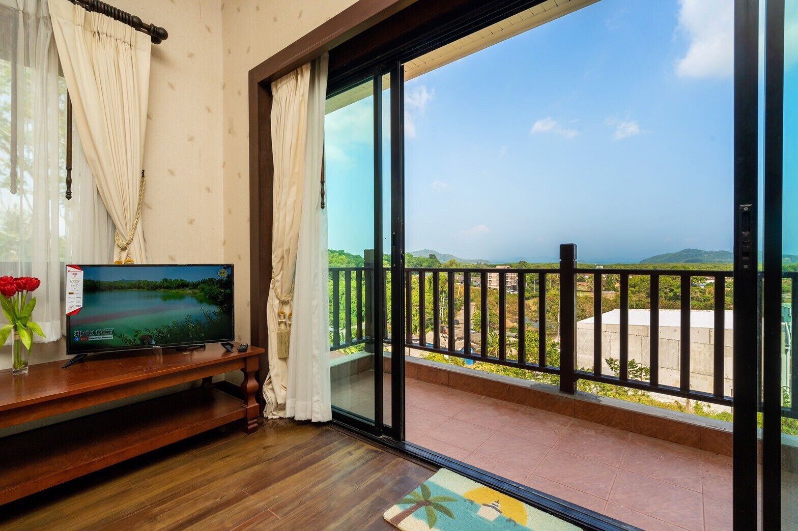 A room with a television on a wooden stand, a glass door leading to a balcony with a view of greenery and hills.