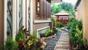 A narrow pathway leading to a traditional gazebo surrounded by lush greenery and a building with large windows and a wooden slatted awning. - Angel Pool Villa Phuket (Ratsada)