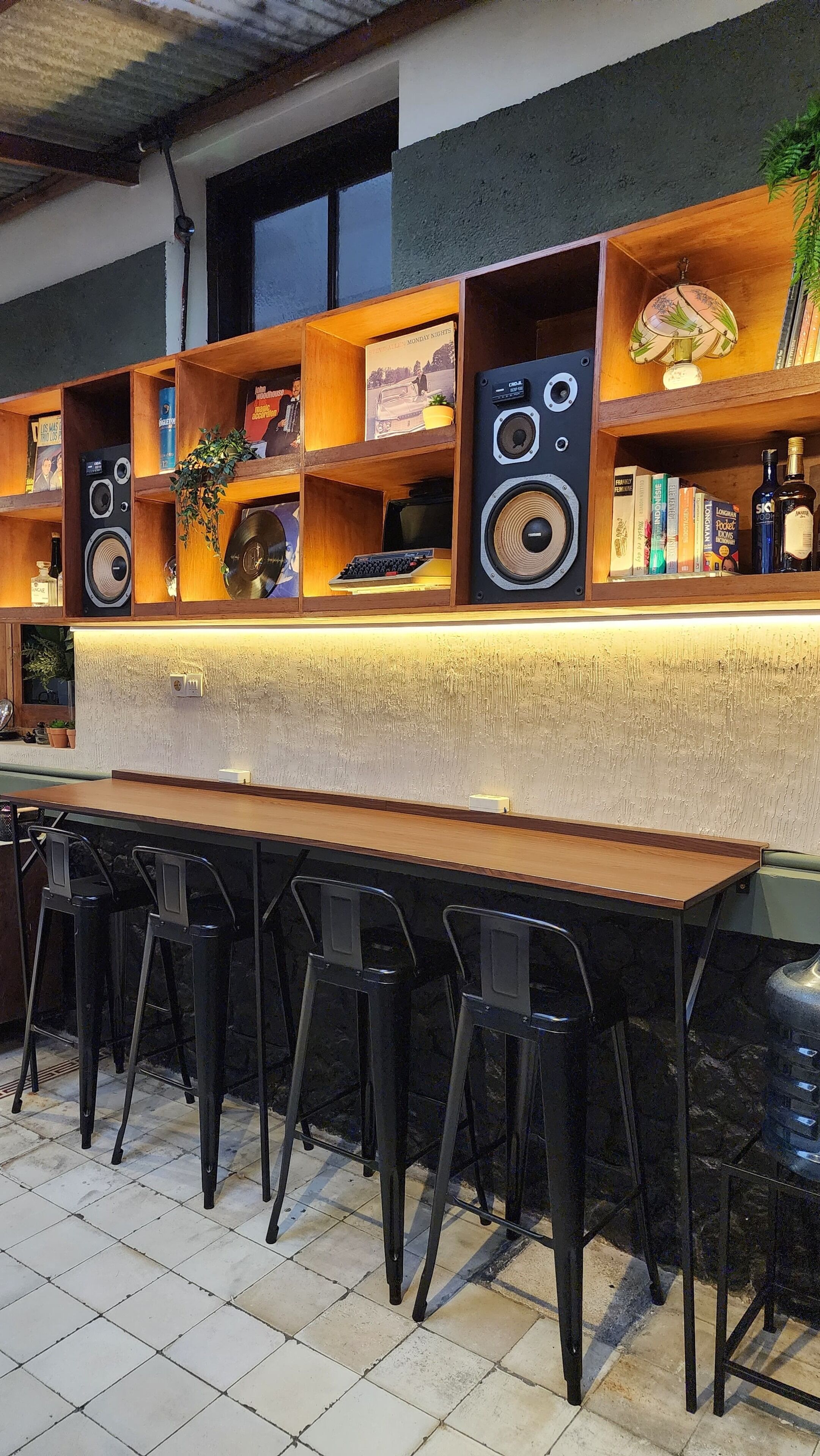 Una zona de bar con una larga barra de madera, taburetes negros y estantes con varios objetos, incluyendo libros, una lámpara y altavoces.