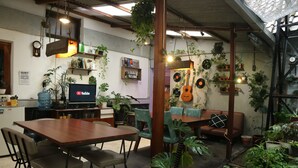 A cozy, well-lit room with a wooden dining table, chairs, and a sofa. There are various potted plants and a guitar wall decoration. - Semeru Hostel Malang - Adults Only (Malang)