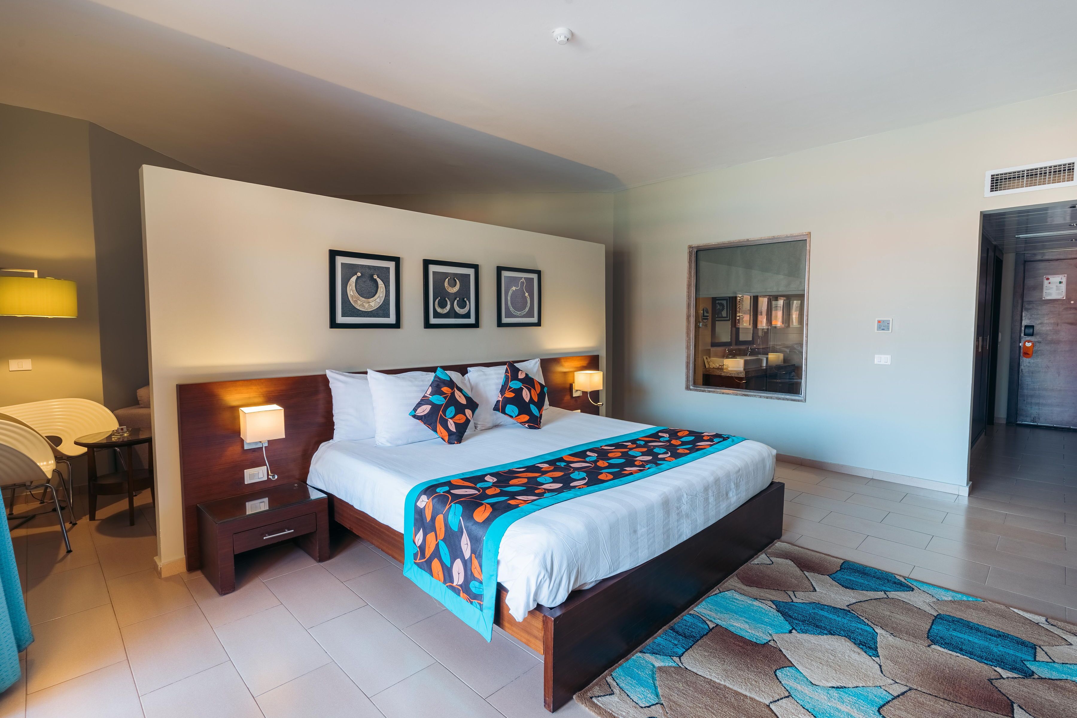 Una habitación de hotel moderna con una cama grande, mesitas de noche con lámparas, una alfombra estampada y cuadros en la pared.