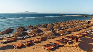 Une plage bordée de rangées de chaises longues et d’ombrelles en chaume, une vue dégagée sur la mer et une montagne lointaine.