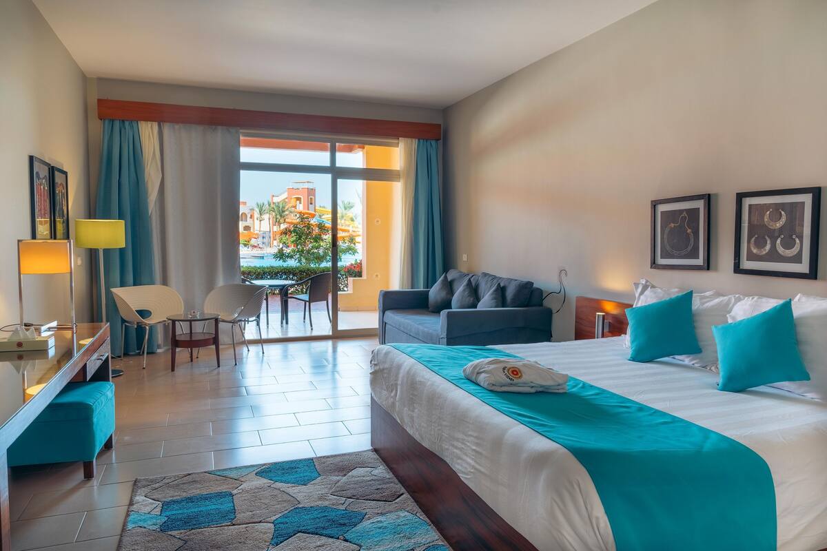 A hotel room with a large bed, a sofa, a desk with a lamp, and a view of the outside.