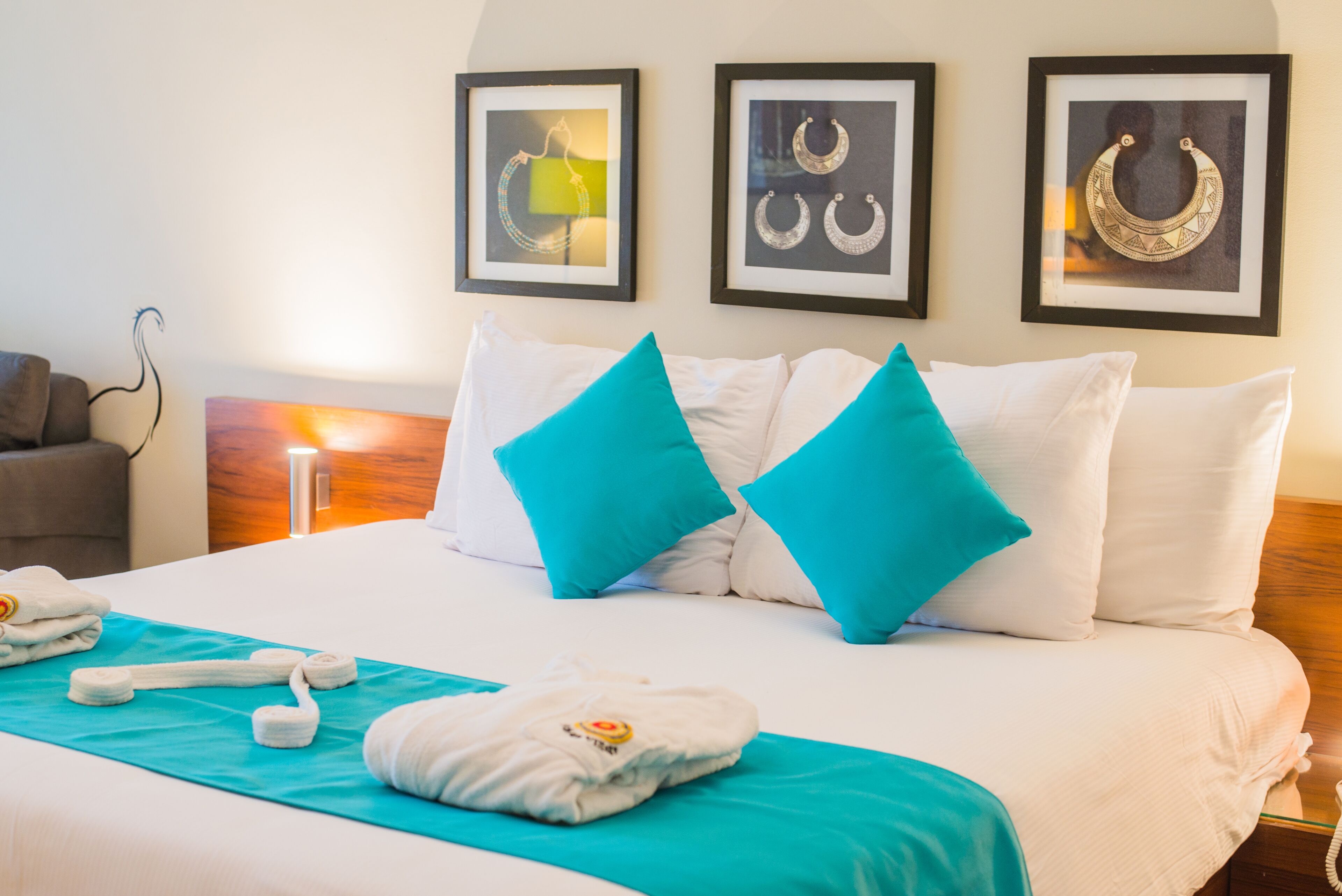 Een hotelkamer met een bed, witte kussens, een blauwgroene deken en een handdoek. Er hangen afbeeldingen in lijstjes aan de muur en het bed heeft een houten hoofdbord.