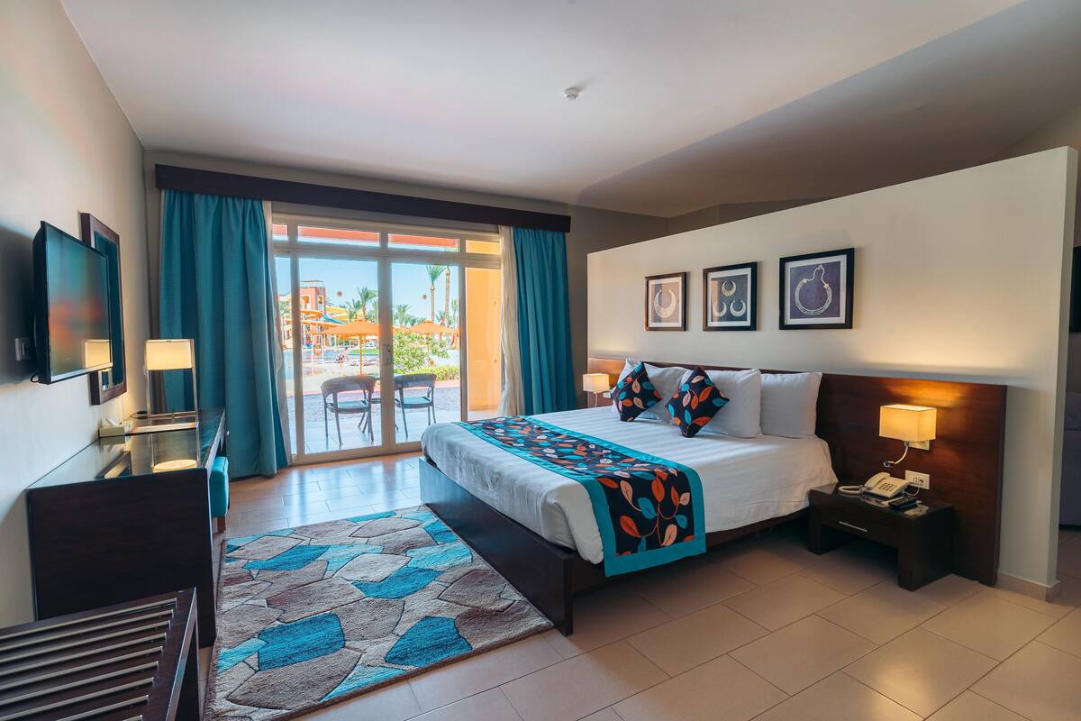 A hotel room with a bed, bedside table, lamp, TV, and a view of the outside.