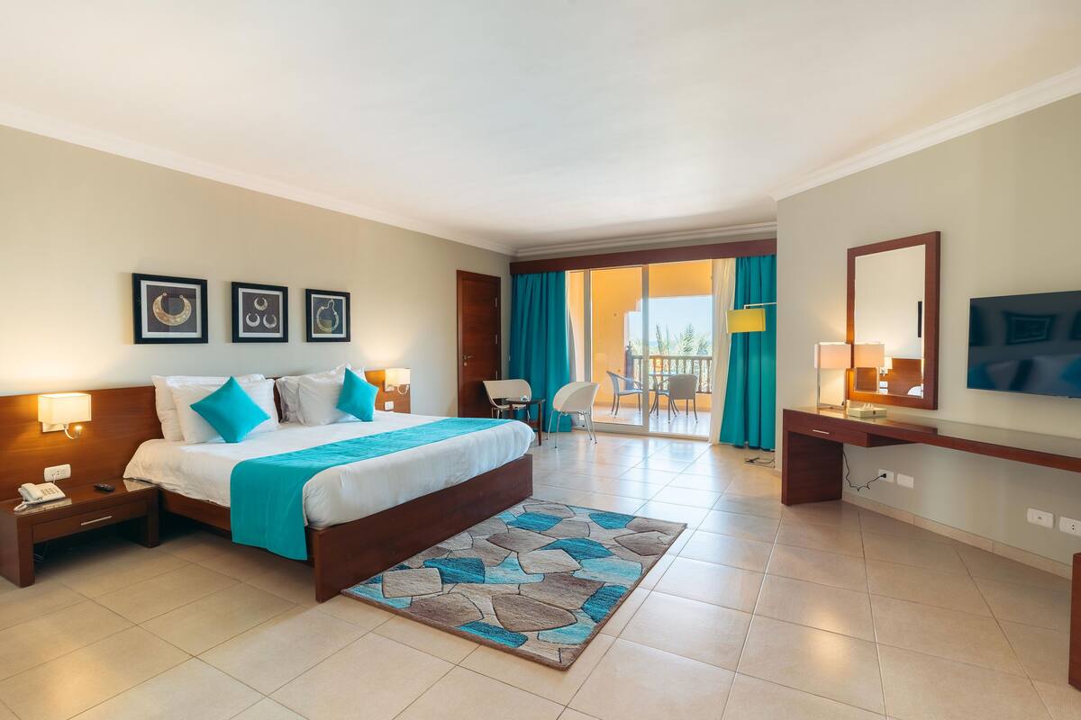 A hotel room with a bed, bedside tables, a desk, a chair, a TV, and a view of the outside.