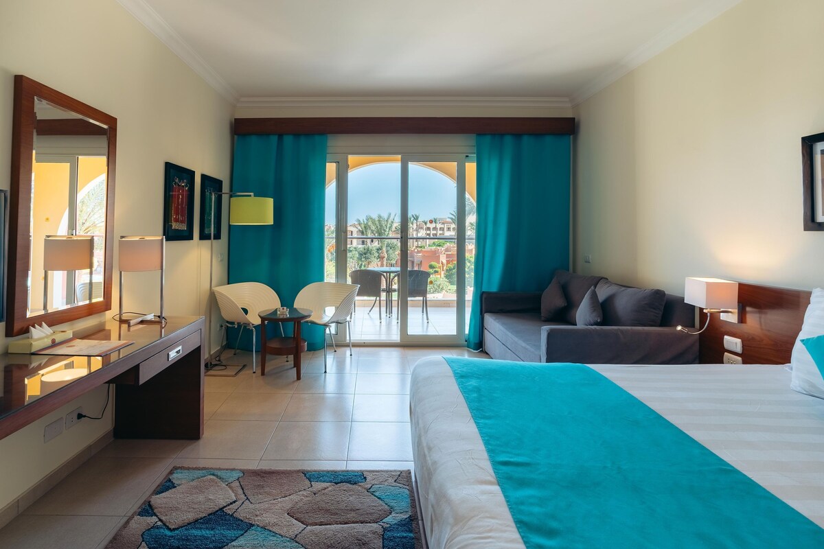 A hotel room with a bed, a desk, a chair, a sofa, a balcony with a view, and a large window.