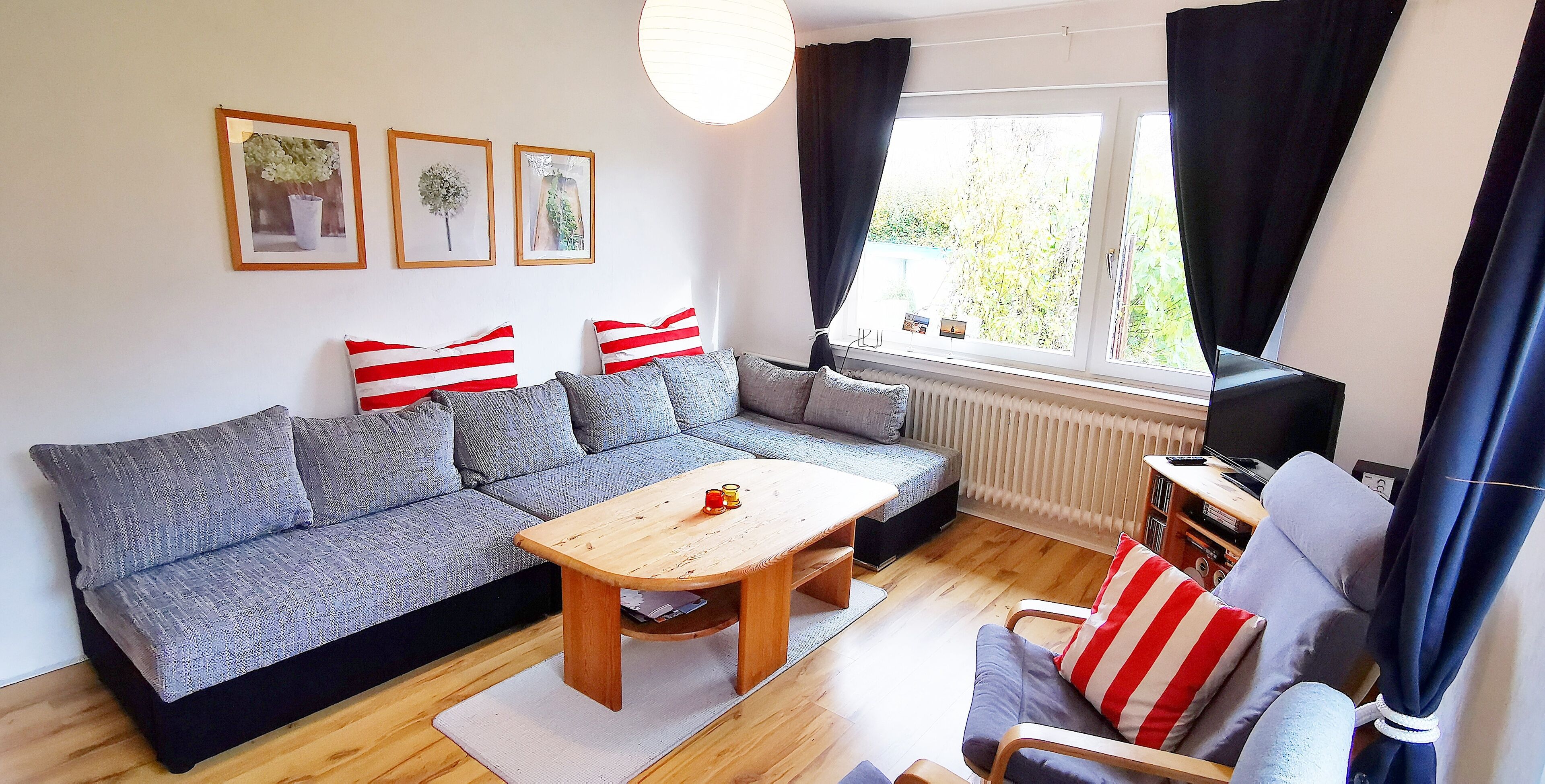 Ein Wohnzimmer mit grauem Sofa, einem Holztisch und einem blauen Sessel mit roten und weißen Streifen-Kissen. An der Wand hängen drei gerahmte Bilder, ein Fenster mit Vorhängen und ein Heizkörper.