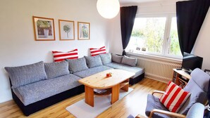Ein Wohnzimmer mit grauem Sofa, einem Holztisch und einem blauen Sessel mit roten und weißen Streifen-Kissen. An der Wand hängen drei gerahmte Bilder, ein Fenster mit Vorhängen und ein Heizkörper.