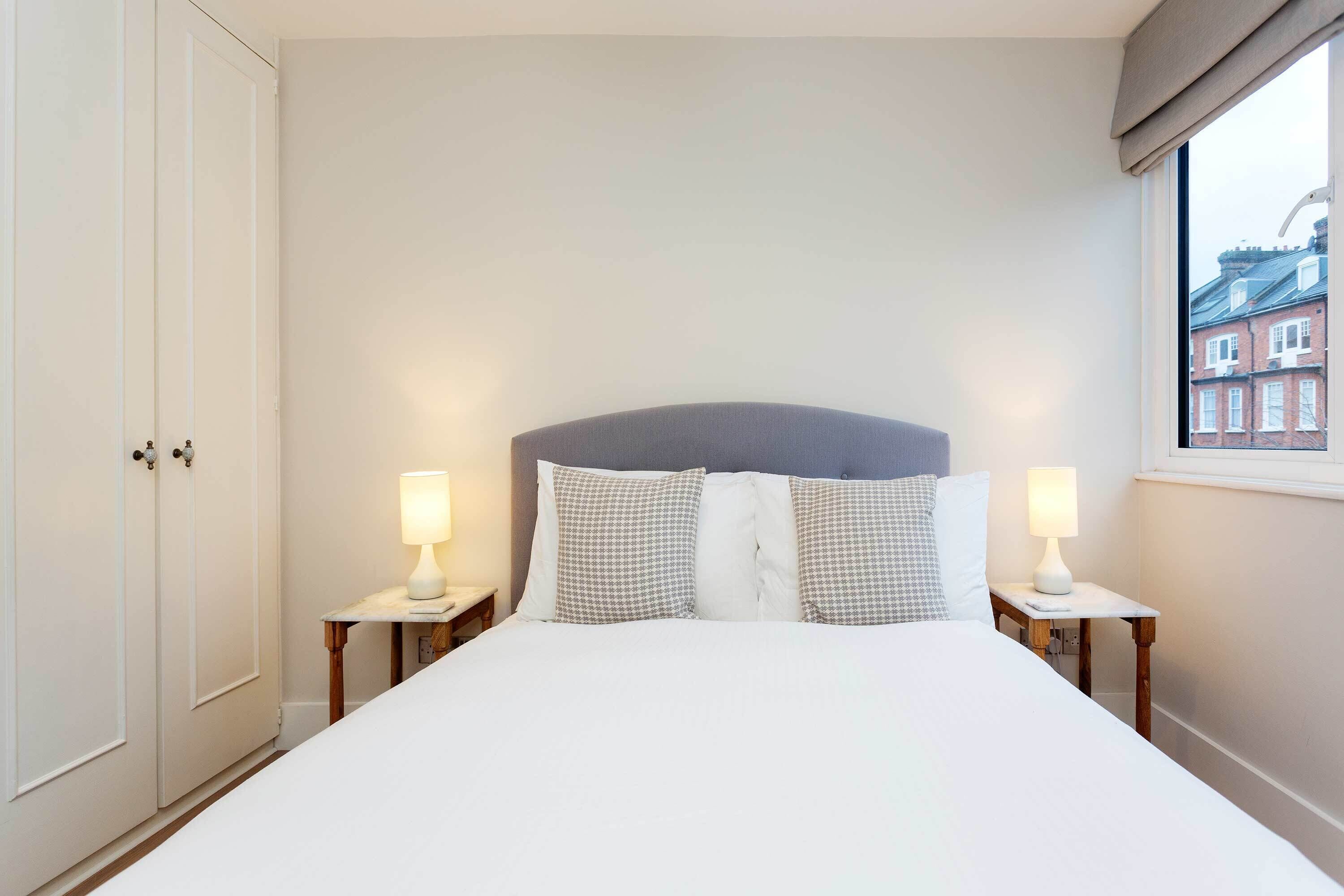 Een opgemaakt bed met witte lakens, twee nachtkastjes met lampen en een raam met uitzicht op gebouwen.