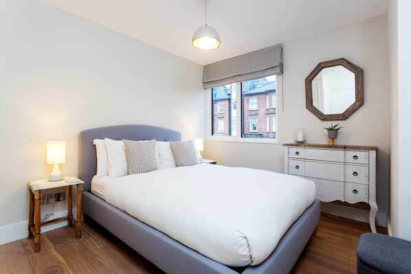 A bedroom with a bed, bedside table, dresser, window, and a hanging mirror. - Calming Belsize Park Retreat, by Veeve (London)
