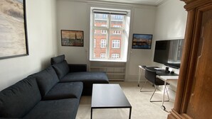 A living room with a dark grey sectional sofa, a small black table, a TV on a stand, and a window with a view of a red brick building. - PSG 23 - Short Stay Apts by Living Suites (Copenhagen)