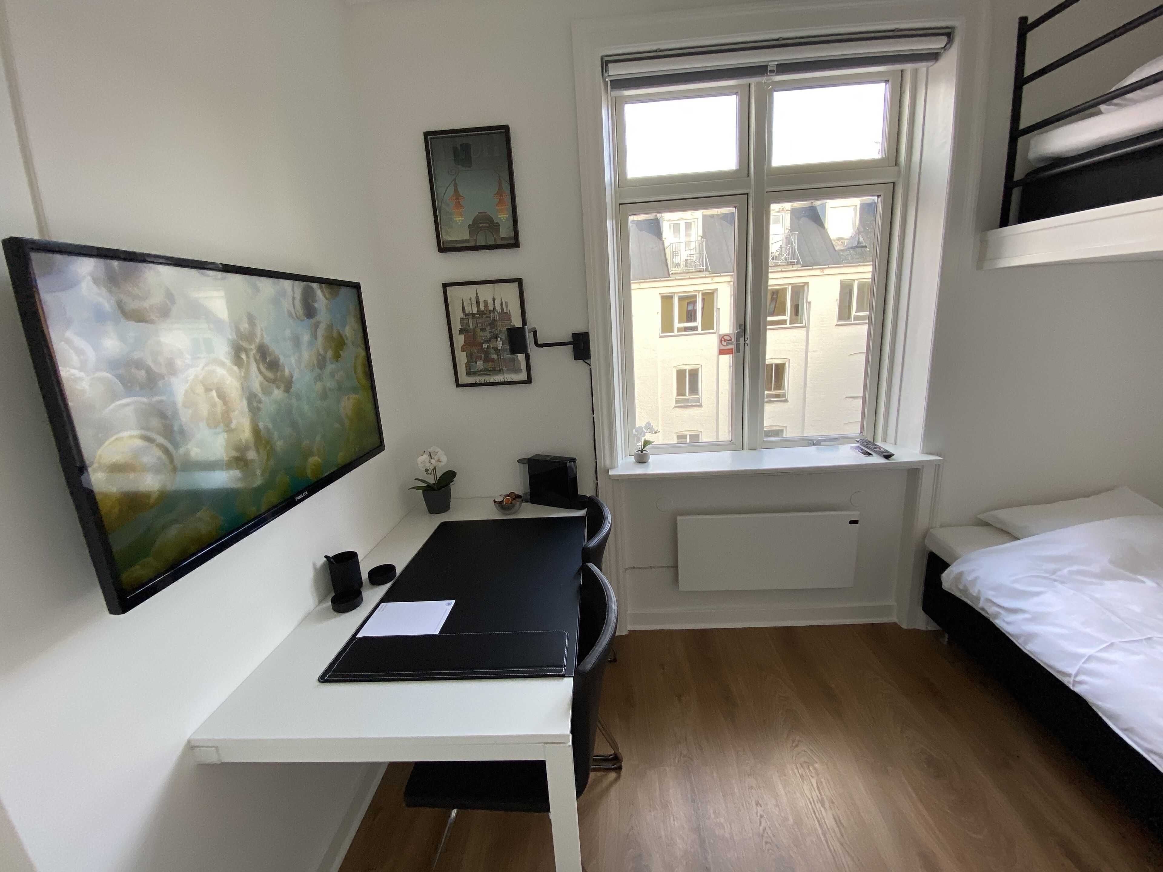 Een kleine, net kamer met een bed, een bureau en een raam met uitzicht op gebouwen.