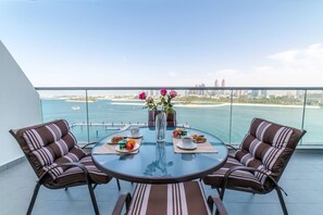 A round glass table with two striped chairs, a vase with pink flowers, and breakfast items on a balcony overlooking a city and water.
