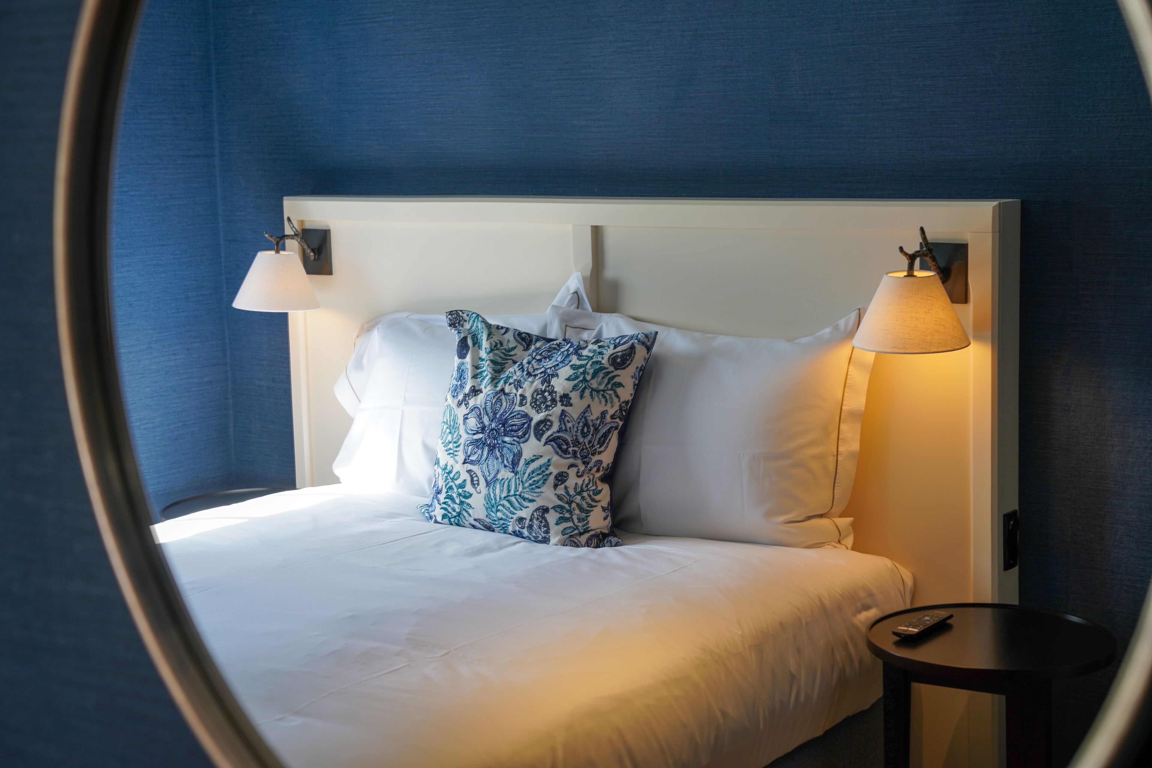 Una cama bien hecha con sábanas blancas y una almohada estampada, una lámpara de noche y una mesita con un control remoto.
