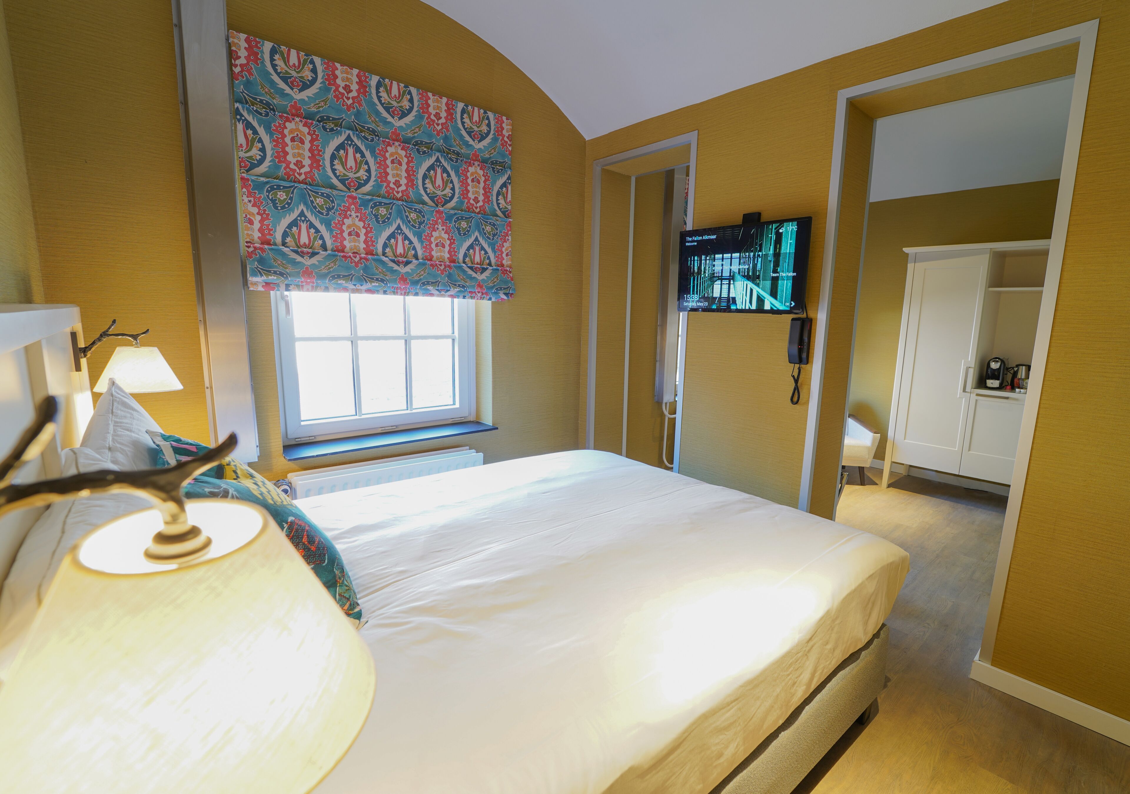 A hotel room with a bed, a TV mounted on the wall, a window with patterned curtains, and a door leading to a bathroom.