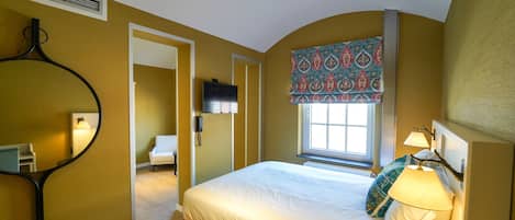 Un dormitorio con cama, una ventana con cortinas estampadas, una lámpara de noche y un televisor montado en la pared.