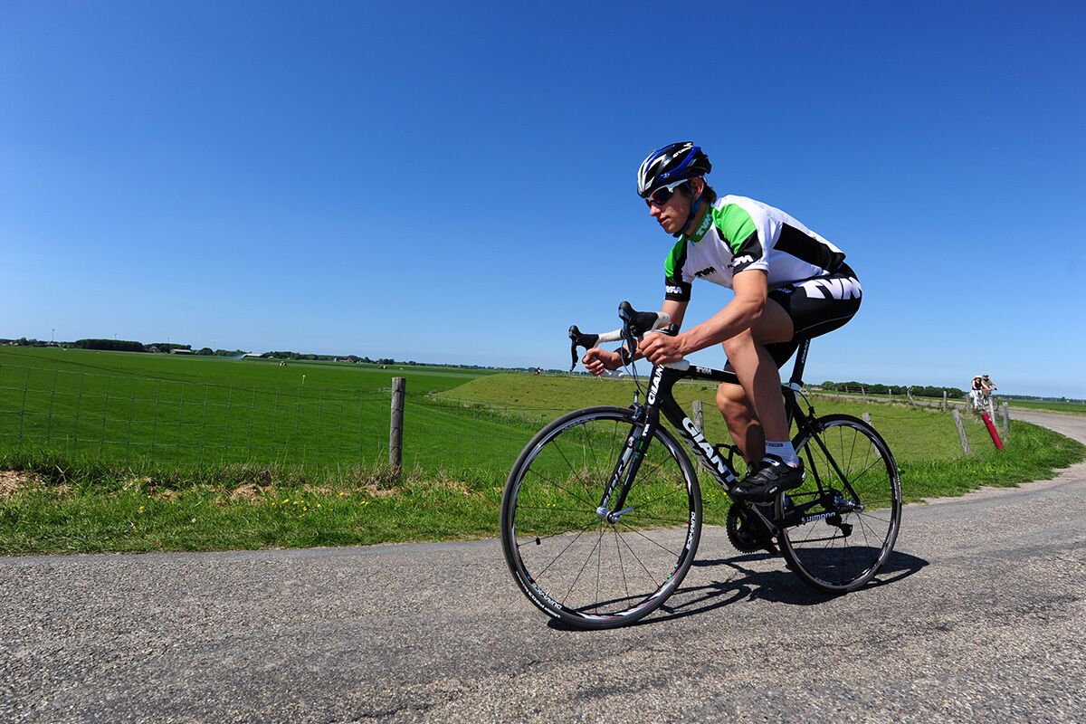 Een fietser op een weg, met een heldere lucht en groene velden op de achtergrond.