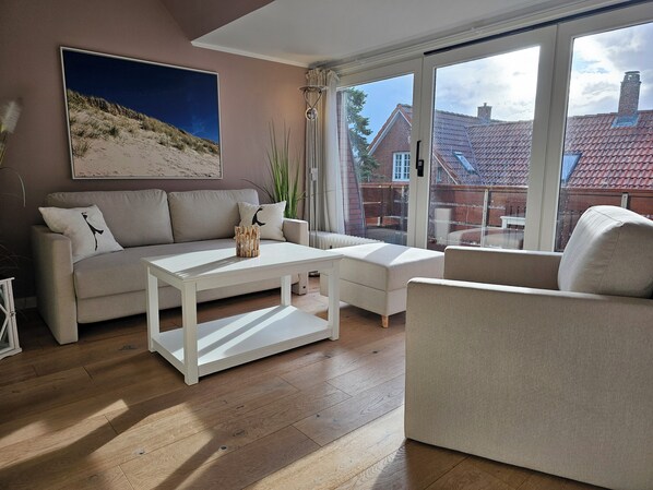 A modern living room with a sofa, a coffee table, and a chair. There is a large window with a view of a house and a garden.