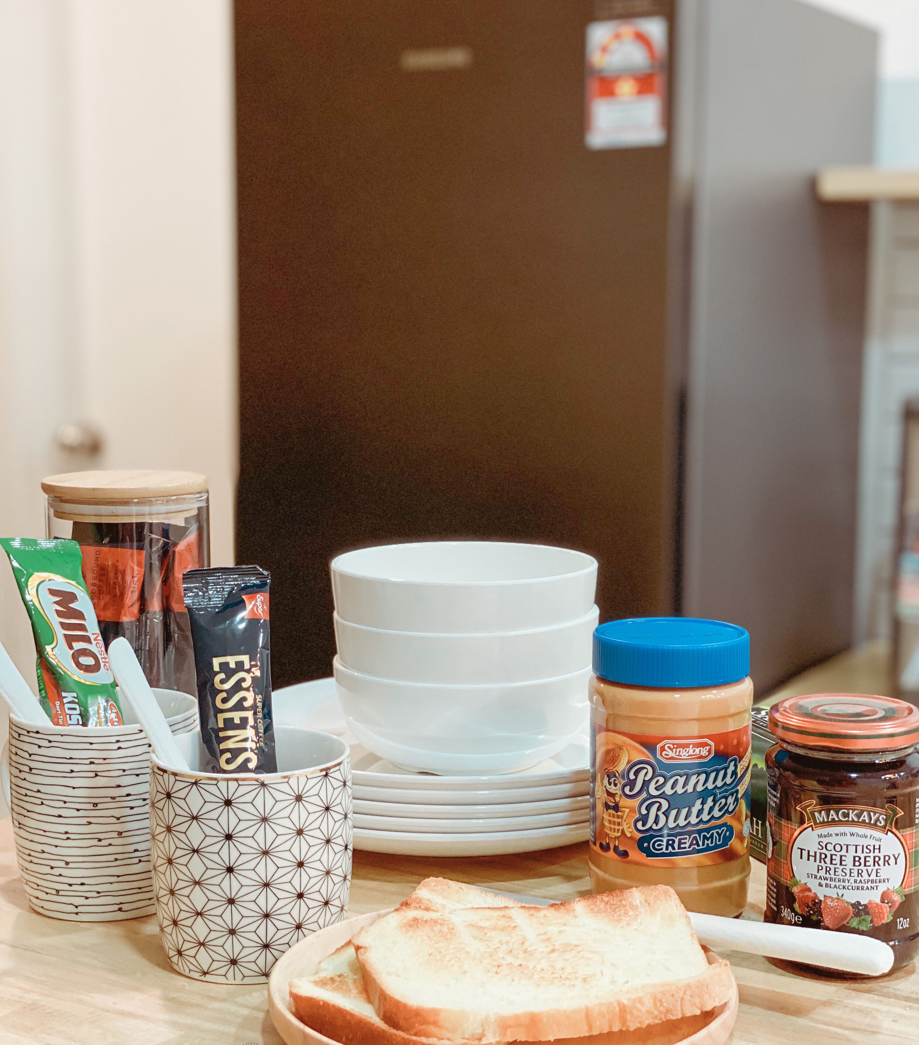 Una encimera de cocina con una pila de platos blancos, un tarro de mantequilla de maní, un tarro de mermelada, un paquete de café y un paquete de té.