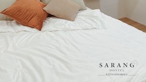 Een net opgemaakt bed met witte lakens en twee kussens, één oranje en één beige. Een wandlamp met houten voet en een macramé wanddecoratie zijn zichtbaar. De kamer heeft een minimalistisch ontwerp met een schoon, wit kleurenschema.
