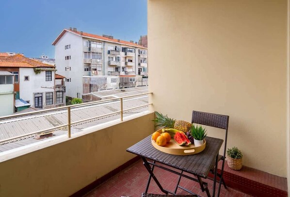 Ein Balkon mit Tisch, gedeckt mit Früchten, und einem Stuhl, mit Blick auf ein Wohngebiet mit Gebäuden.