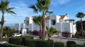 Un bâtiment moderne de couleur blanche, doté de nombreux balcons, entouré de palmiers et de haies bien entretenues.