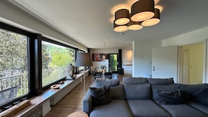 Un salon moderne avec un canapé gris, une table ronde en bois et une grande fenêtre donnant sur un balcon.
