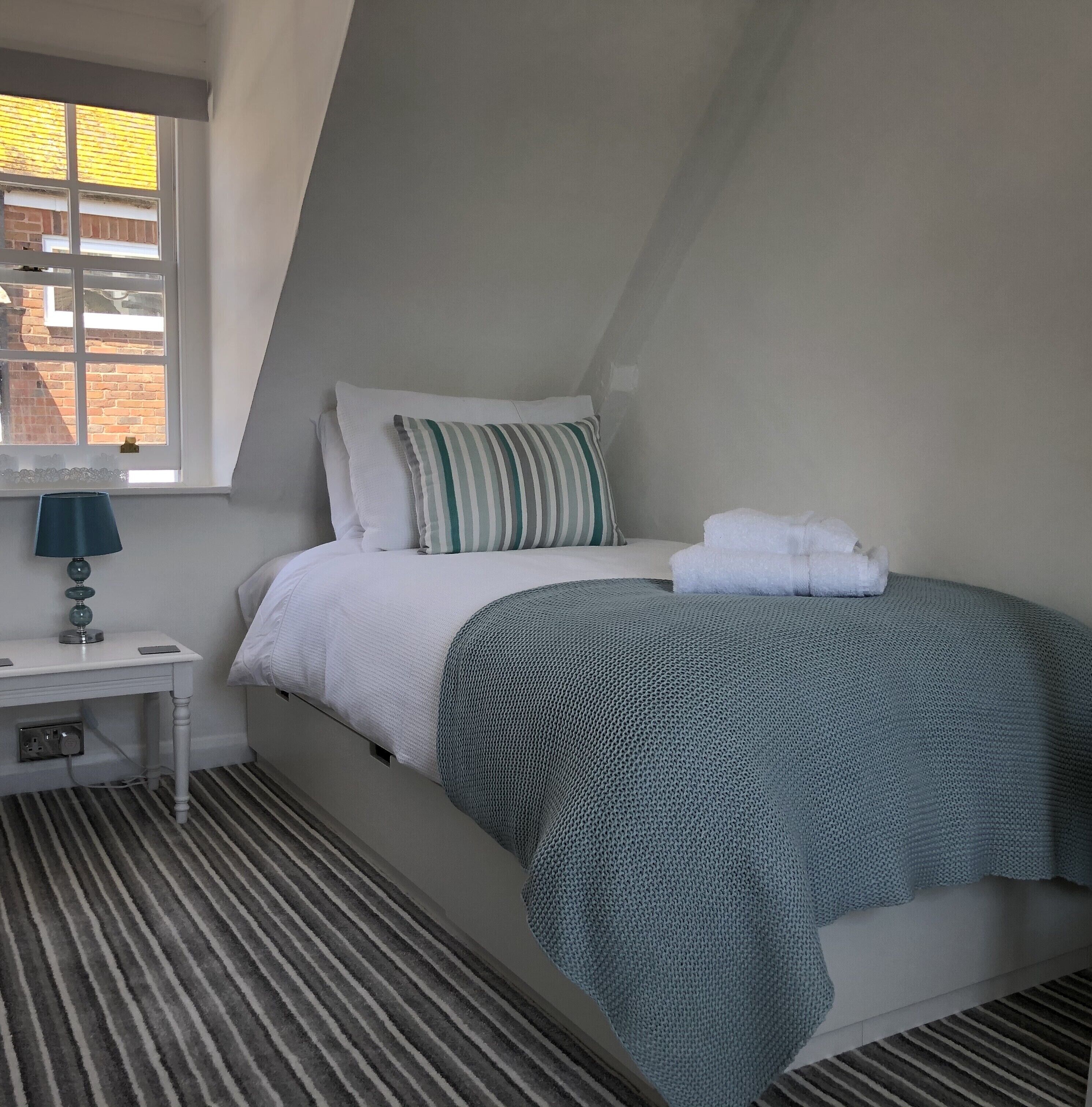Un dormitorio con cama, mesita de noche con lámpara, ventana y alfombra a rayas.