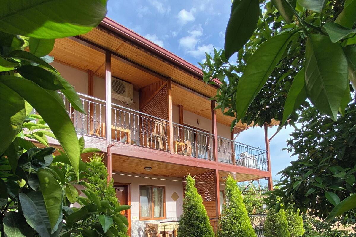 A two-story building with a balcony, surrounded by greenery and a stone pathway.