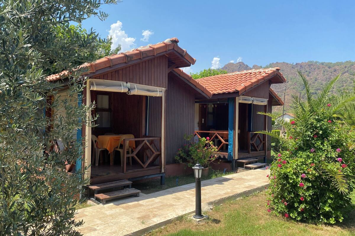 A rustic wooden house with a tiled roof, a covered porch, and a small garden with flowering plants.