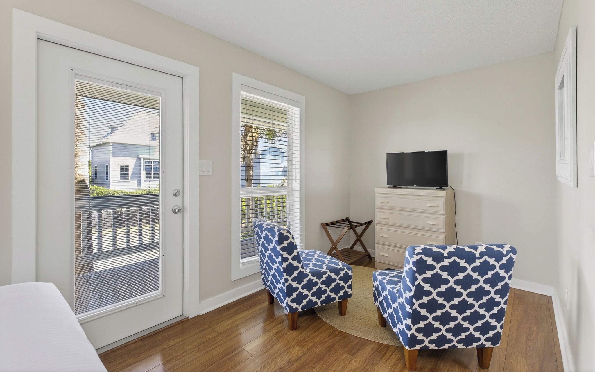 A room with a sliding glass door, two patterned armchairs, a small wooden table, a dresser with a TV on top, and a view of a house through the door.