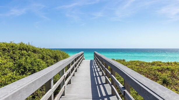 Une promenade en bois qui mène à une plage bordée d’une eau turquoise cristalline et sous un ciel dégagé.