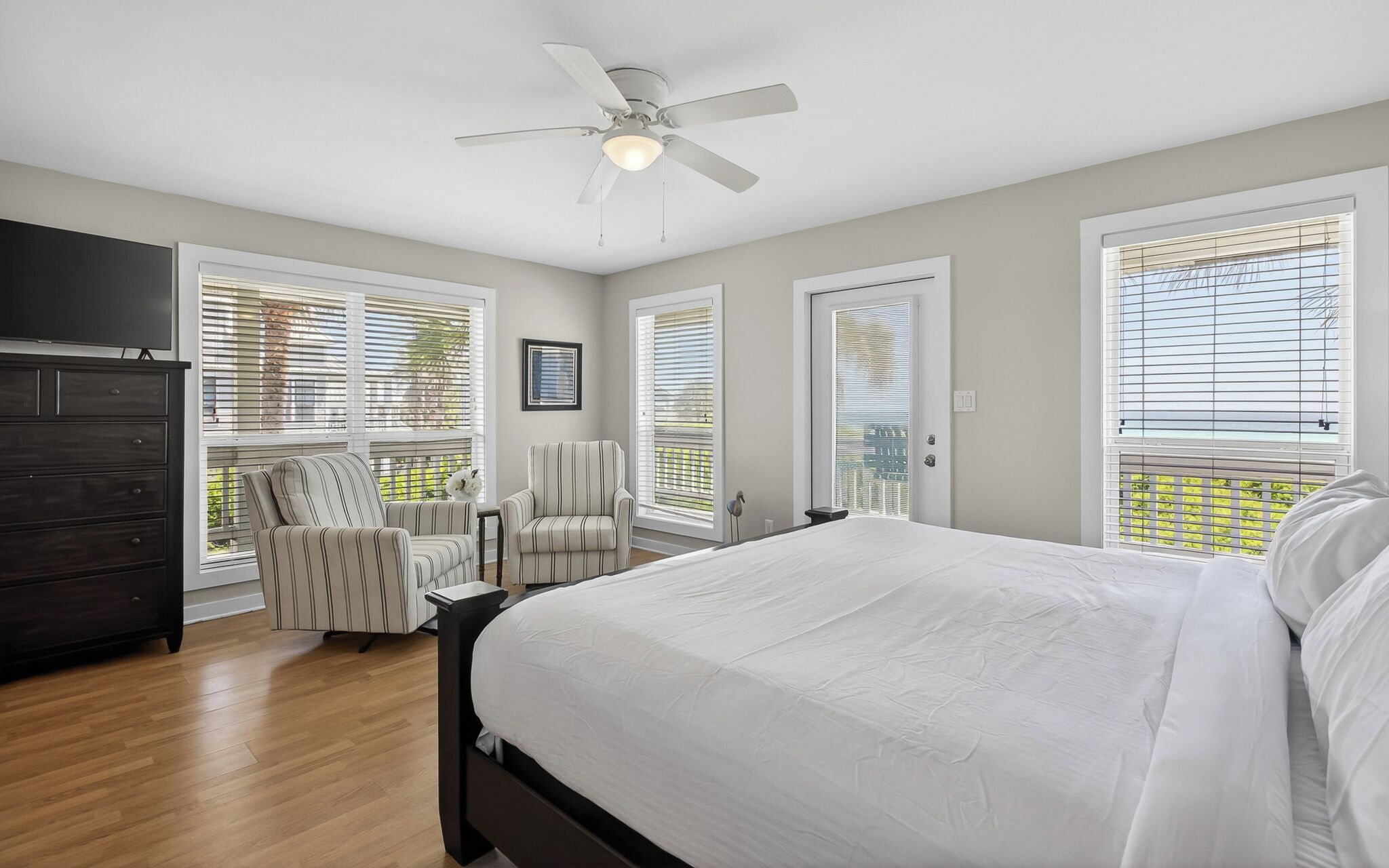 Une chambre à coucher comprenant un lit, une commode, une chaise, un ventilateur de plafond et de grandes fenêtres équipées de stores.