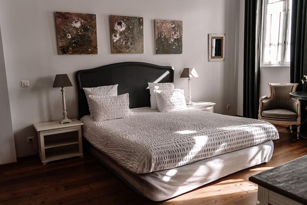 A neatly made bed with a white textured bedspread and pillows, flanked by bedside tables with lamps, in a room with wooden flooring and artwork on the walls.